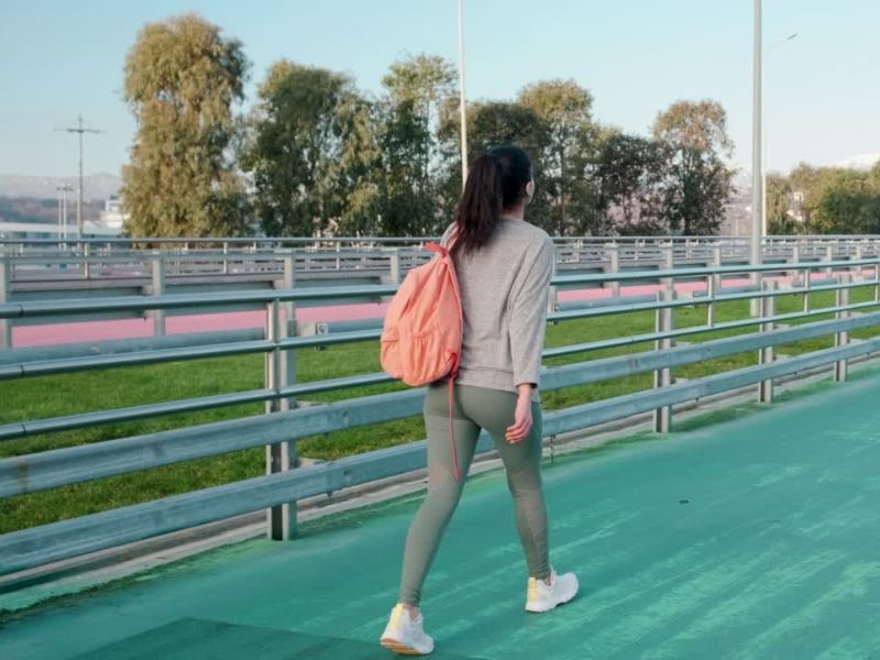 Devojak duge crne kose sportski obučena, sa rancem na leđima šeta stazom za trčanje