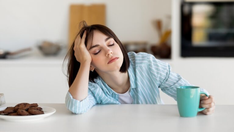 Žena sedi za stolom sa šoljom kafe i deluje iscrpljeno zbog manjka sna